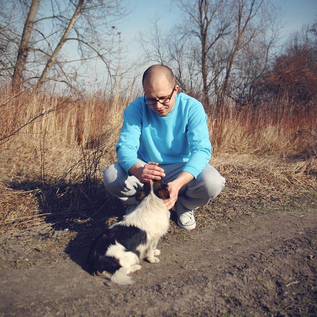 Zdjęcie kolorowe. W tle zarośla i sucha trawa. Na pierwszym planie mężczyzna w okularach i niebieskim swetrze kuca aby pogłaskać psa.