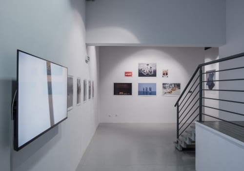 Zdjęcie sali wystawienniczej wewnątrz CSW Łaźnia, podczas trwania wystawy "Widzialne głosy. Młoda fotografia koreańska dzisiaj!". Na jednej ze ścian zawieszony jest telewizor, na którym wyświetlana jest praca wideo. Fotografie są wyeksponowane na białych ścianach.