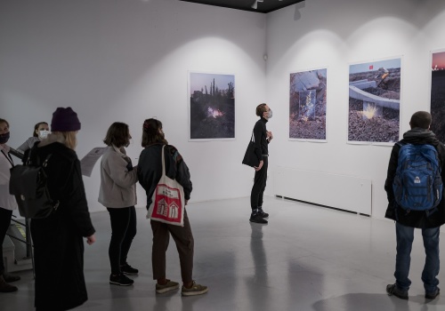 Fotografia widowni wydarzenia "Widzialne głosy. Młoda fotografia koreańska dzisiaj!". Zdjęcie wykonane w przestrzeni wystawienniczej, wewnątrz CSW Łaźnia. Goście wydarzenia mają założone maseczki ochronne i z uwagą przyglądają się zaprezentowanym dziełom.