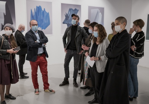 Fotografia widowni wydarzenia "Widzialne głosy. Młoda fotografia koreańska dzisiaj!". Zdjęcie wykonane w przestrzeni wystawienniczej, wewnątrz CSW Łaźnia. Goście wydarzenia posiadają założone maseczki ochronne. W tle tłumu ukazują się zaprezentowane dzieła, wyeksponowane na białych ścianach przestrzeni wystawienniczej.