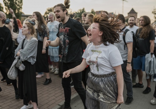 Fotografia pozioma pokazująca grupę uczestników koncertu.  Na pierwszym planie kobieta i mężczyzna tańczą i wykrzykują teksty piosenek. Ich włosy są rozwiane, na twarzach wypisana ekstaza. Jest letni słoneczny dzień. Na drugim planie, ponad głowami fanów, widać  zarys panoramy Głównego Miasta.