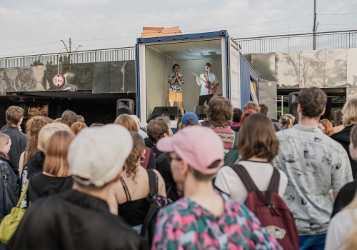 Fotografia pozioma pokazująca na pierwszym planie od tyłu tłum uczestników koncertu. Na drugim planie widać przeszkloną witrynę kontenera LKW Gallery, a wewnątrz artyści Franek Warzywa i Młody B grający koncert. Na trzecim planie widać wiadukt obity lustrzanymi płytami.