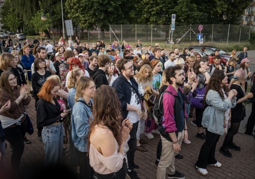 Fotografia pozioma pokazująca grupę uczestników koncertu. Są to młodzi ludzie. Niektóry mają kolorowe włosy, niektóry klaszczą. Jest letni słoneczny dzień. Na drugim planie widać  drzewa i ulicę.