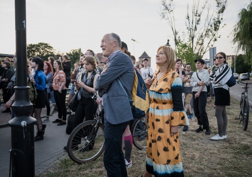  Fotografia pozioma pokazująca grupę uczestników koncertu. Na pierwszym planie dwoje ludzi w wieku ok. 60-70 lat z radością i zainteresowaniem patrzących w kierunku artystów. Na drugim planie więcej młodych fanów i osoby z rowerami. Jest letni słoneczny dzień. W dalszym planie widać  drzewa.