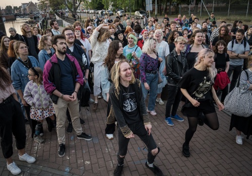 Fotografia pozioma pokazująca tłum uczestników koncertu. Są to przede wszystkim młodzi ludzie. Niektóry mają kolorowe włosy. Niektóry tańczą i śpiewają. W dalszym planie widać szpaler zielonych drzew, ulicę, kanał Motławy, kamienice Głównego Miasta i most Stągiewny.