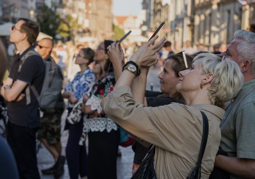 Widok na grupę osób biorących udział w spacerze z oprowadzeniem po ulicy Długiej, które fotografują smartfonami fasady kamienic. W tle widok na wieżę Ratusza Głównego Miasta.