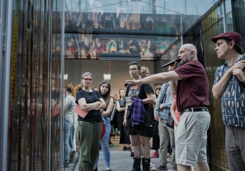 Artysta Marek Płóciennik z mikrofonem wskazujący ręką dzieło artystyczne z zawieszonych klisz fotograficznych, we wnętrzu Kunsztu Wodnego, siedziby Instytutu Kultury Miejskiej. Wokół tłum osób słuchających artystę.