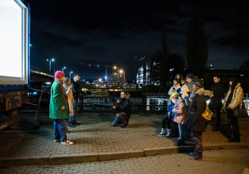 Zdjęcie. Grupa ludzi stojąca bokiem do kamery, zwróconych twarzami w stronę  przeszklonej witryny kontenera LKW Gallery w ciężarówce. Między nimi a ciężarówką stoją artystka Natalia Szerszeń, kuratorka Anna Szynwelska i tłumacz PJM, którzy opowiadają o wystawie. Na drugim planie widać kanał Motławy i nocny miejski pejzaż.  