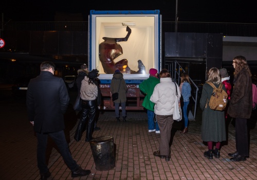 Zdjęcie. Rozproszona  grupa ludzi stojących tyłem do kamery, zwróconych twarzami w stronę  przeszklonej witryny kontenera LKW Gallery w ciężarówce, gdzie znajduje się rzeźba przedstawiająca upadającego konia.